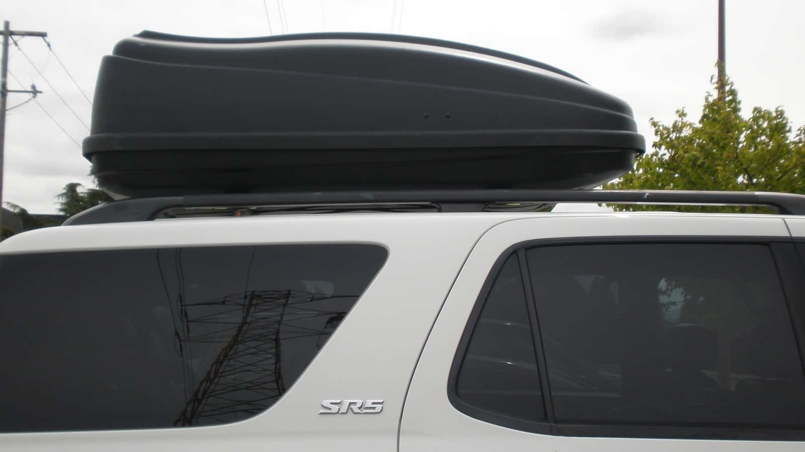 A black hard-shell cargo box mounted on the roof-rack assembly of a white Toyota SUV.