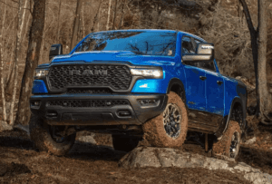 A bold blue RAM 1500 pickup truck navigating an off-road trail, with its wheels covered in mud and front tire elevated on a rock, surrounded by a wooded forest in early spring.