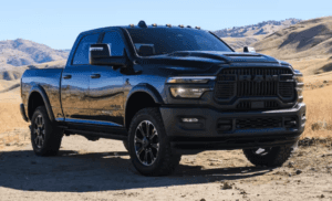 A black RAM 2500 heavy-duty pickup truck parked on a dirt path in a dry, mountainous landscape, showcasing its rugged front grille, raised hood, and aggressive off-road stance under bright sunlight.