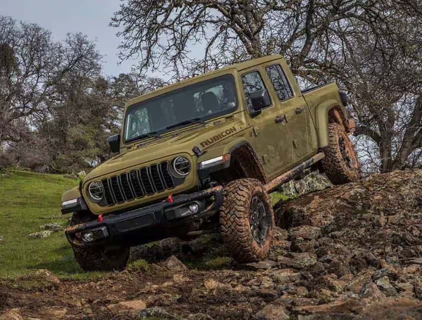An olive green Jeep Gladiator Rubicon tackles a rocky incline on a rugged off-road trail, with its suspension flexed and tires gripping the uneven terrain. The scene is set in a hilly area with sparse trees and overcast skies, highlighting the truck’s trail-rated capability.