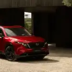 2026 Mazda CX-5 near Uniondale NY in Soul Red Crystal with black wheels and trim parked on gravel in front of a dark wood architectural structure