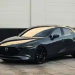 2026 Mazda3 Sedan near Uniondale NY in dark gray with black wheels parked on concrete lot in front of a white and black industrial building