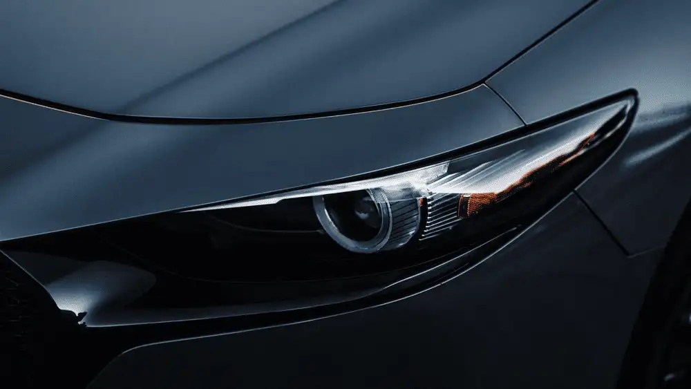 2026 Mazda3 Sedan near Uniondale NY close-up of sleek projector headlight with LED ring on dark gray hood in dramatic low lighting