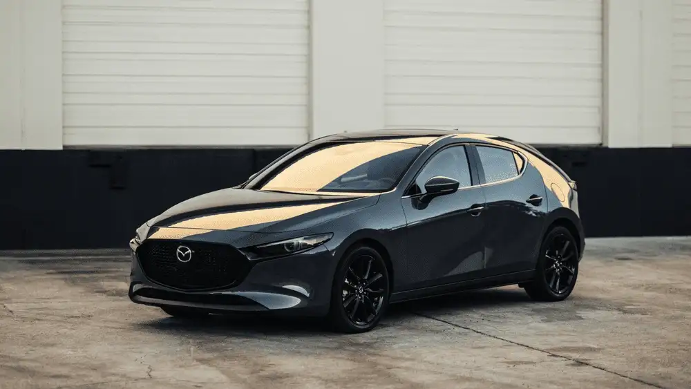 2026 Mazda3 Sedan near Uniondale NY in dark gray with black wheels parked on concrete lot in front of a white and black industrial building