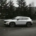 2026 Mazda CX-50 ground clearance built for Queens exploration shown in white driving in profile on open road surrounded by tall pine forest in black and white