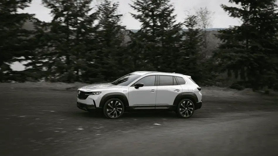 2026 Mazda CX-50 ground clearance built for Queens exploration shown in white driving in profile on open road surrounded by tall pine forest in black and white
