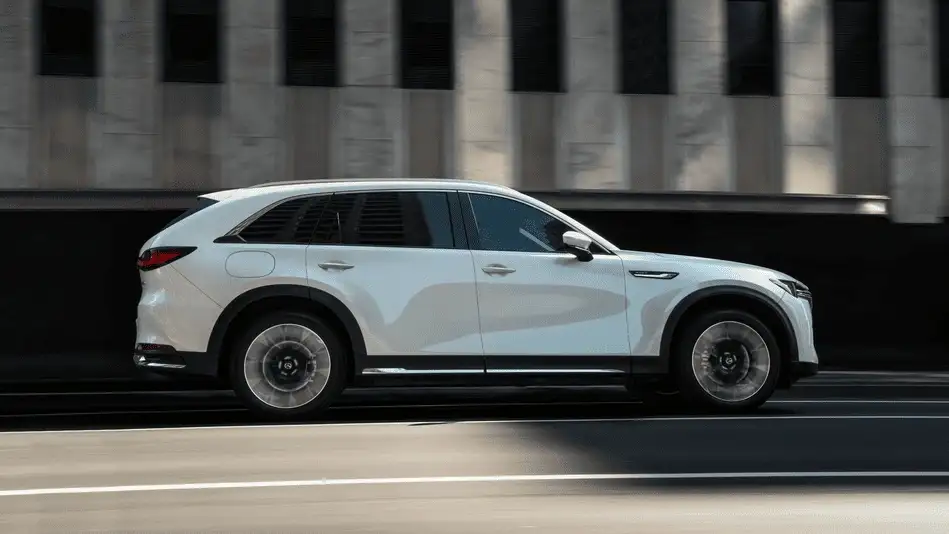 2026 Mazda CX-90 Plug-In Hybrid out to Corona NY in Queens in white driving at speed along urban highway with motion-blurred concrete building in background