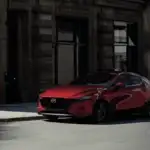 2026 Mazda3 Hatchback delivering fun weekend drives around Corona NY in Soul Red parked on a shadowy urban street beside a historic stone building