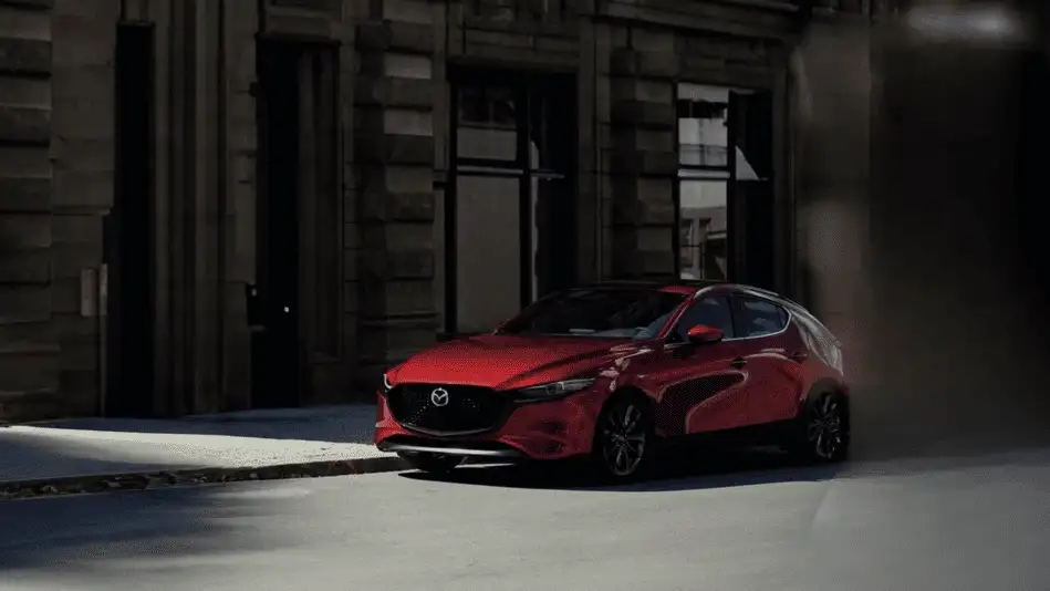 2026 Mazda3 Hatchback delivering fun weekend drives around Corona NY in Soul Red parked on a shadowy urban street beside a historic stone building