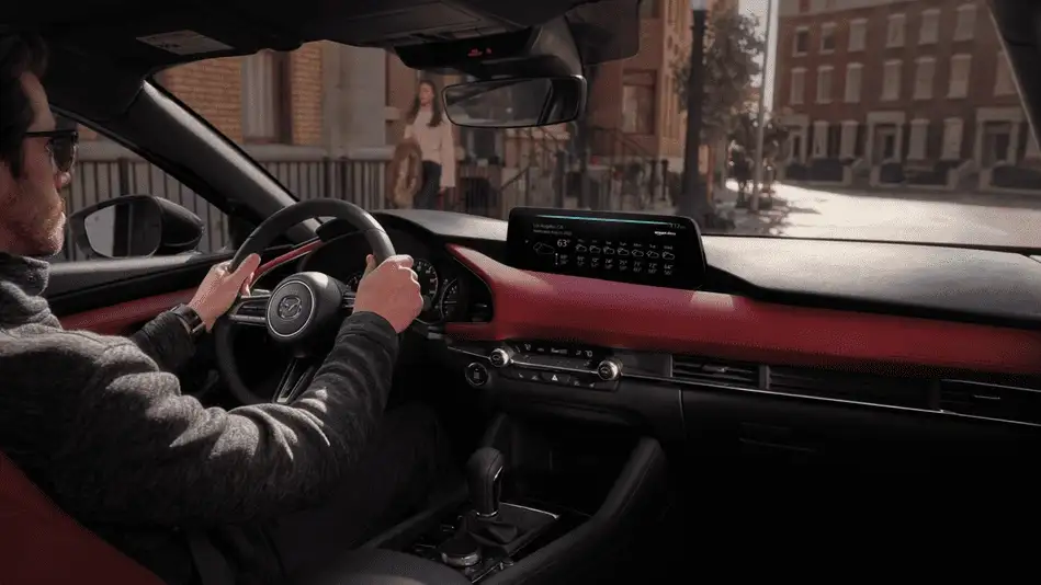 2026 Mazda3 Hatchback out to Corona NY in Queens driver gripping steering wheel with red dashboard trim, infotainment screen showing weather, and city street through windshield