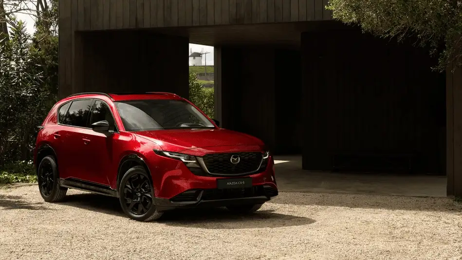 2026 Mazda CX-5 in Soul Red with black wheels on gravel driveway framed by dark wood architecture, a top choice versus the Subaru Forester around Corona NY