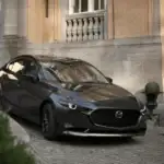 2026 Mazda3 Sedan in dark gray parked on cobblestone driveway in front of a warmly lit stone estate, representing refined driving through Queens city streets