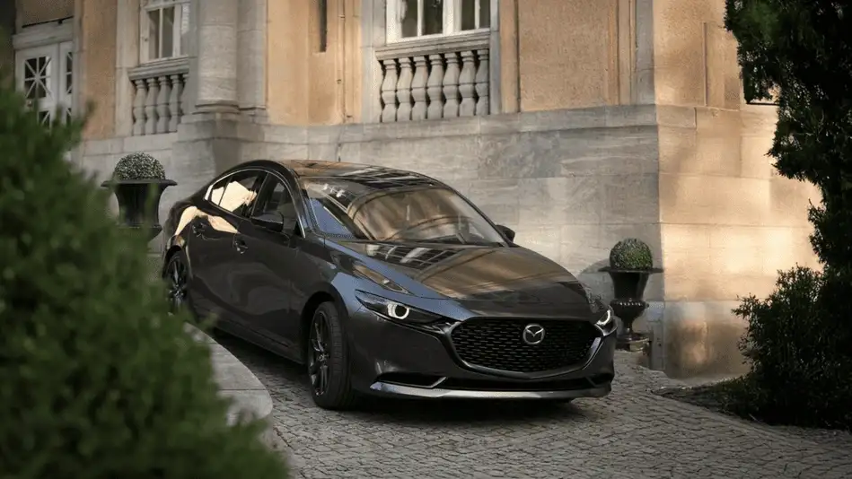2026 Mazda3 Sedan in dark gray parked on cobblestone driveway in front of a warmly lit stone estate, representing refined driving through Queens city streets