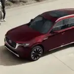 2026 Mazda CX-90 Plug-In Hybrid in deep red viewed from elevated angle showing panoramic roof and large alloy wheels parked on open plaza with two pedestrians nearby