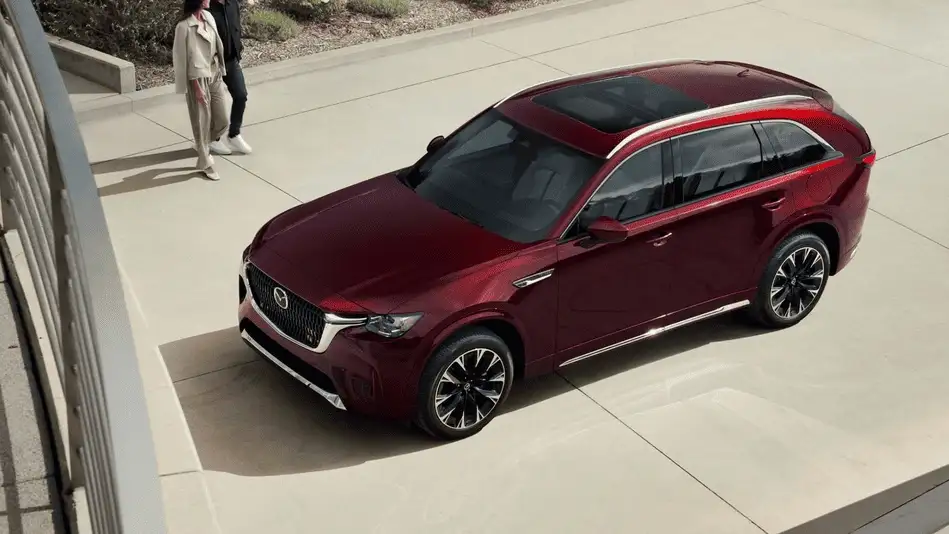 2026 Mazda CX-90 Plug-In Hybrid in deep red viewed from elevated angle showing panoramic roof and large alloy wheels parked on open plaza with two pedestrians nearby