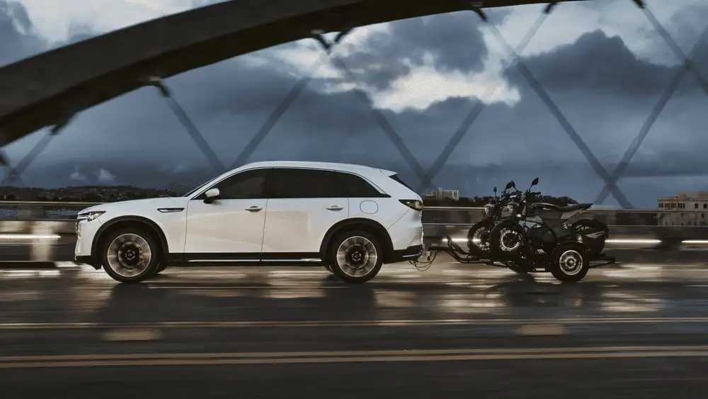 2026 Mazda CX-90 Plug-In Hybrid close to Uniondale NY in white towing a motorcycle trailer across a bridge at dusk with dramatic cloudy sky in background