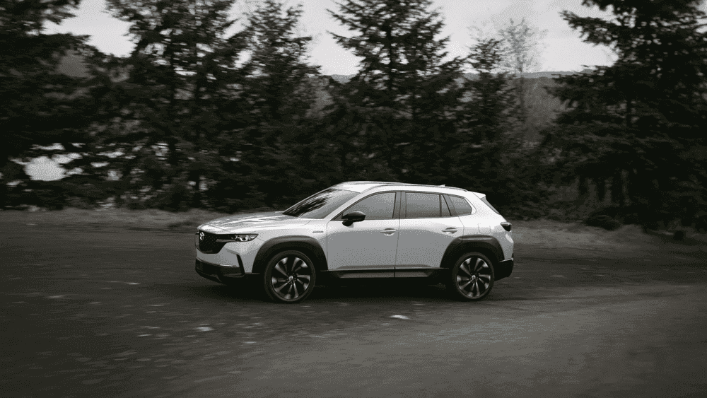 2026 Mazda CX-50 Hybrid near Hempstead NY, white SUV driving in profile on open road with pine forest in background, black and white photo