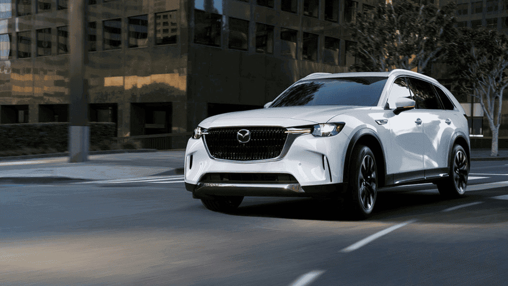2026 Mazda CX-90 near Hempstead NY, white SUV driving through a city intersection with glass office buildings in the background