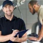 smiling man with clipboard standing in front of mechanic working on car