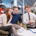couple looking at laptop computer with salesman in new vehicle showroom
