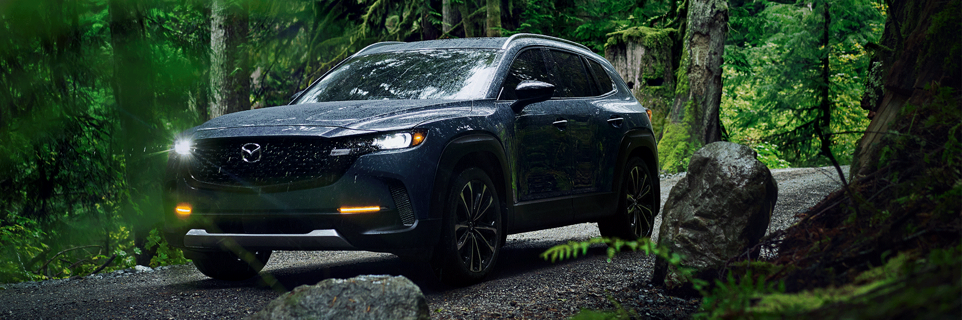 Lancaster Mazda is a Mazda Dealership near Reading, PA | Front Facing Balck 2023 Mazda CX-50 Facing Left Driving through Forest on Rocky Road