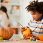 Smiling creative child leaning on counter with gouaches and making design on pumpkin