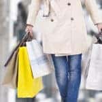A picture of a person from the waist down, who is wearing a light tan trench coat and skinny jeans. They are holding several Christmas shopping bags