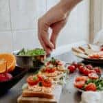 A counter full of vegetables with a vegetarian toast meal on display. A white hand is in the shot to sprinkle salt on the toast.