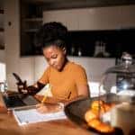Young woman learning online with laptop and phone, she is taking notes in a notebook