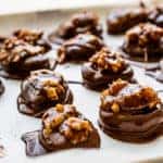 A tray of freshly made chocolate turtle candies. The chocolate is still wet and shiny and there are walnuts on top of each candy mound. There are 16 candy mounds shown.