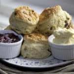 Several scone stacked on a porcelain plate with ramekins filled with raspberry jam and clotted cream.
