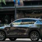 Silver Mazda CX-5 parked on the street in front of a concrete building with some greenery and pedestrians