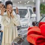 Black man surprising his wife with a used car gift wrapped in a red bow, covering her eyes for the big reveal