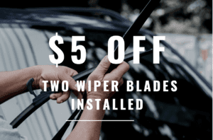 There is a photograph of a car windshield with a reflection on it. In front of the windshield you see two hands of a lighter skin holding two windshield wiper blades. On top of the photograph there is text saying "$5 of two wiper blades installed" in white lettering. 