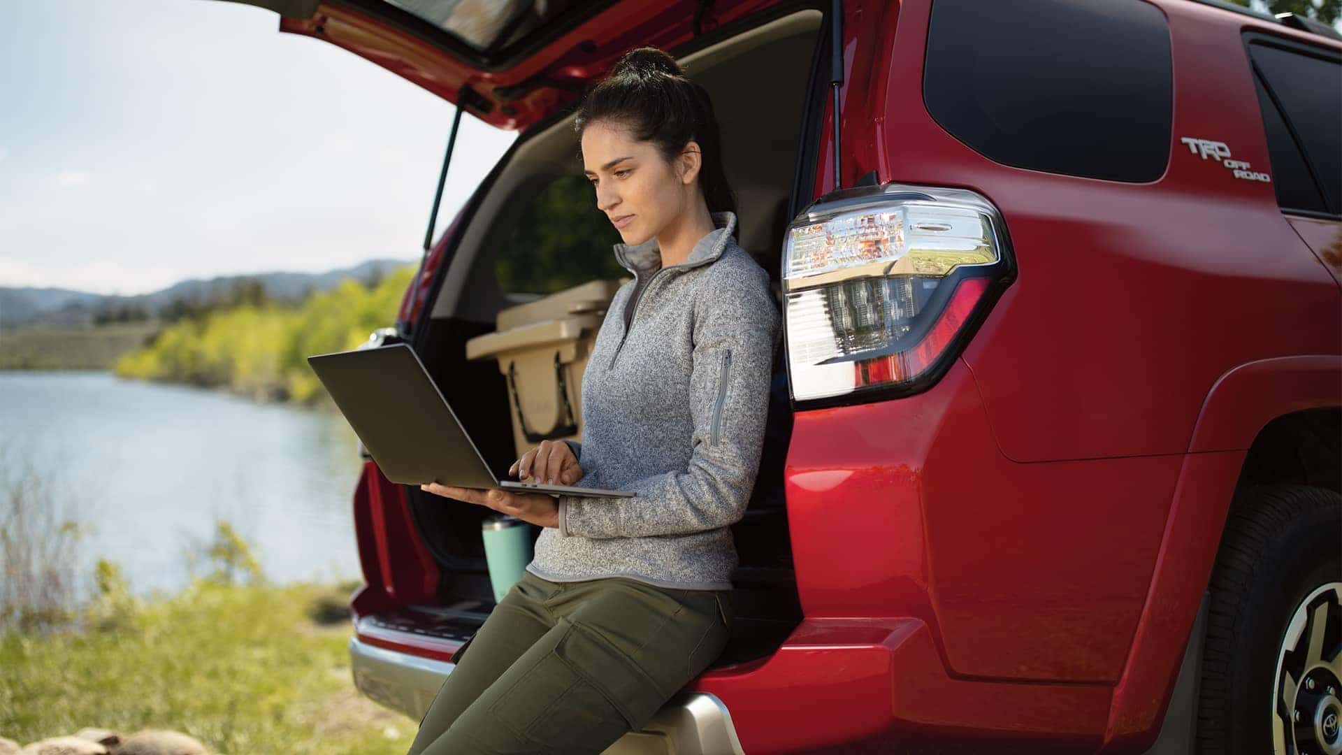 woman on computer sitting in trunk of car near water