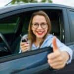 Young happy woman sitting in a car with the keys in her hand and thumb up - concept of buying a used car or a rental car